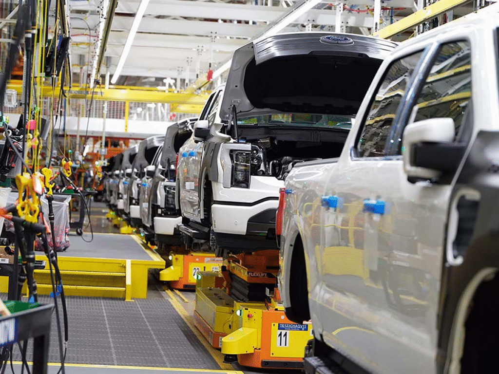 White Ford trucks on an assembly line in a modern manufacturing facility, with open hoods revealing engines, automated guided vehicles positioned underneath, and industrial equipment and yellow safety barriers visible throughout the production floor.