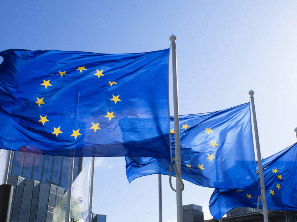 European Union flags flying on tall poles outside modern office buildings on a clear, sunny day