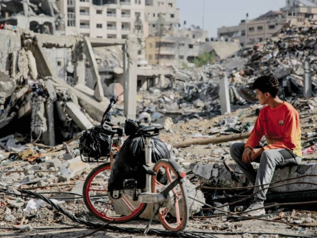 A boy sits beside a bicycle amid collapsed buildings and debris in a heavily damaged area of Gaza.
