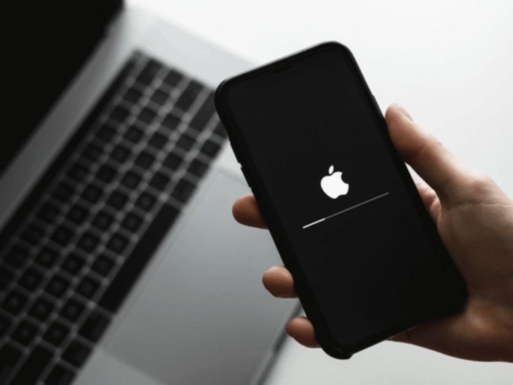An Apple iPhone displaying the Apple logo during startup, held in a hand beside an open laptop.