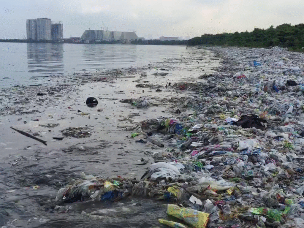 A shoreline heavily covered with plastic waste and debris, with water filled with scattered trash and distant buildings visible across the bay.